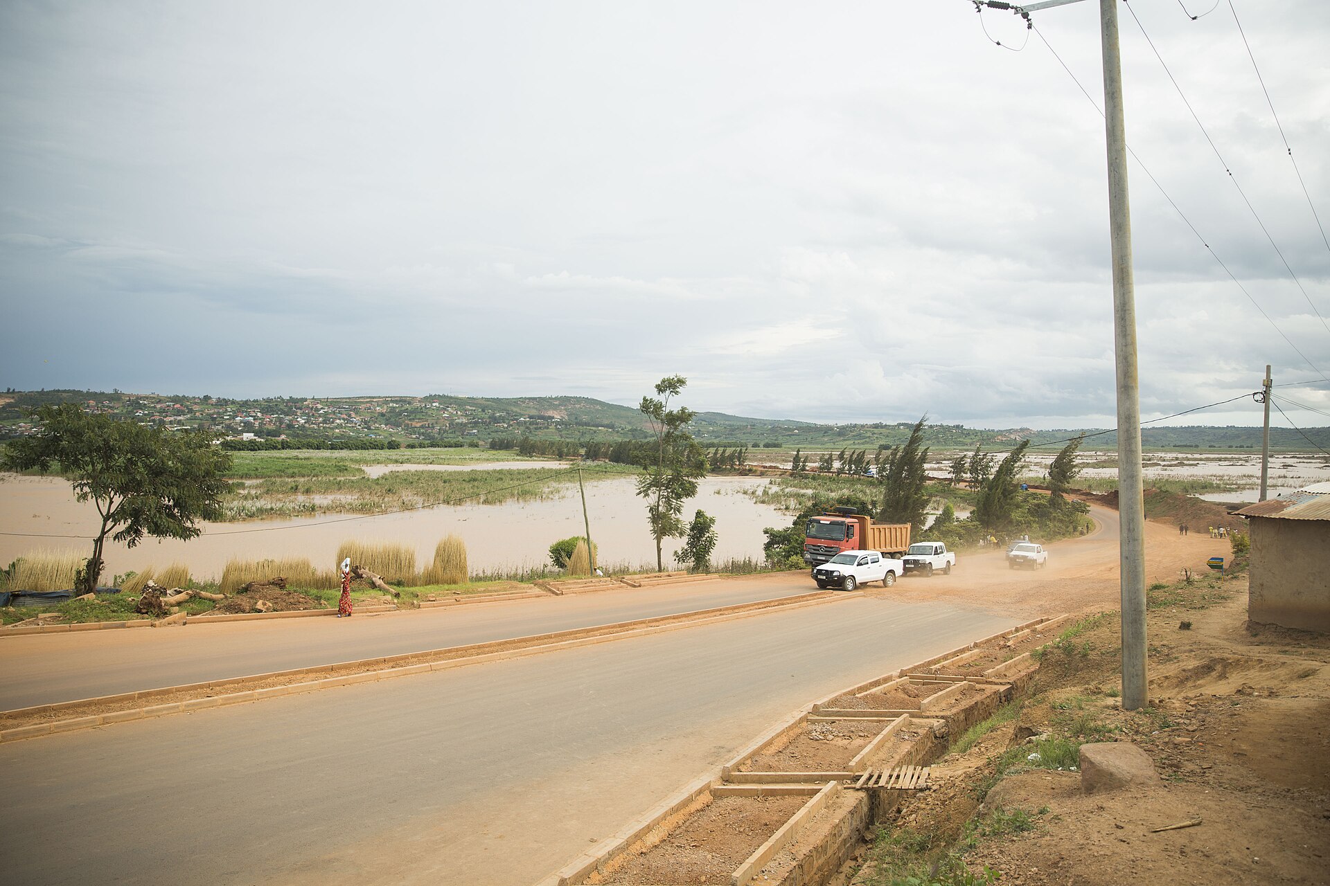 A road beside flooded lowland and low green hills under a grey sky, paved in the foreground and dusty gravel beyond, with an orange lorry and several white pickup trucks driving along it