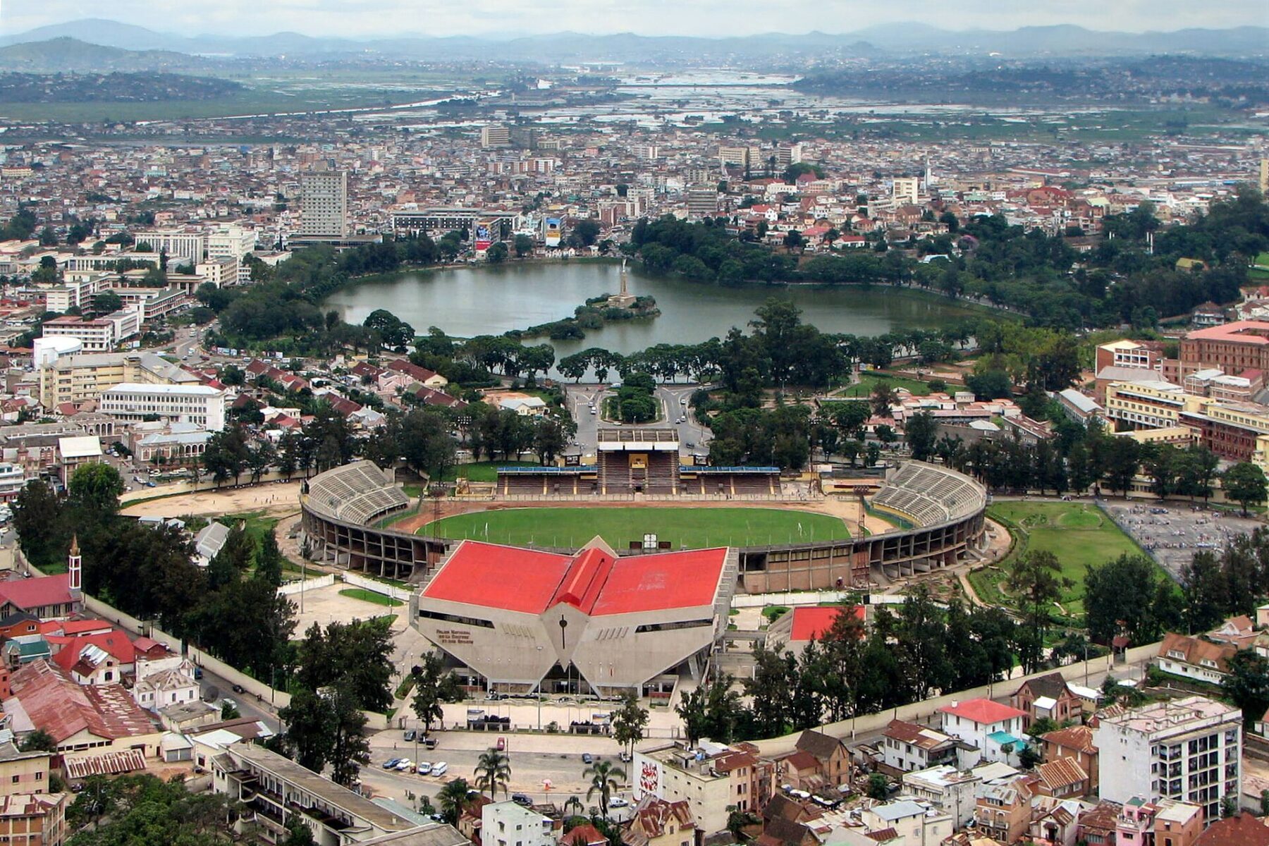 Aerial view of Antananarivo with a lake and island monument, a stadium with a green pitch and a red roofed building in the foreground