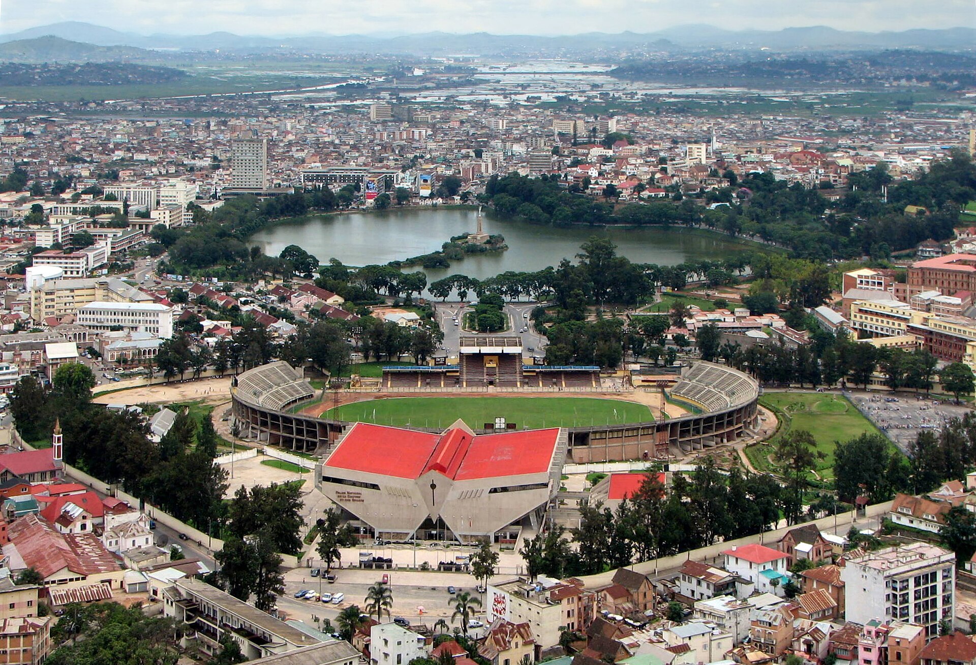 Aerial view of Antananarivo with a lake and island monument, a stadium with a green pitch and a red roofed building in the foreground