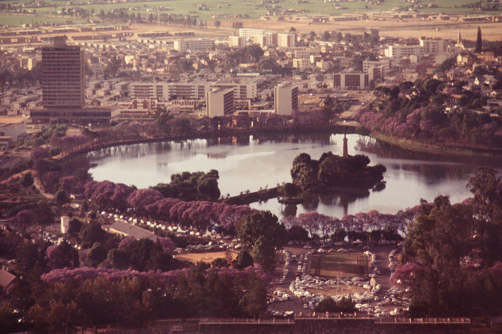 Faded colour aerial photograph of Lake Anosy with a small island and obelisk, purple flowering trees and office blocks beyond