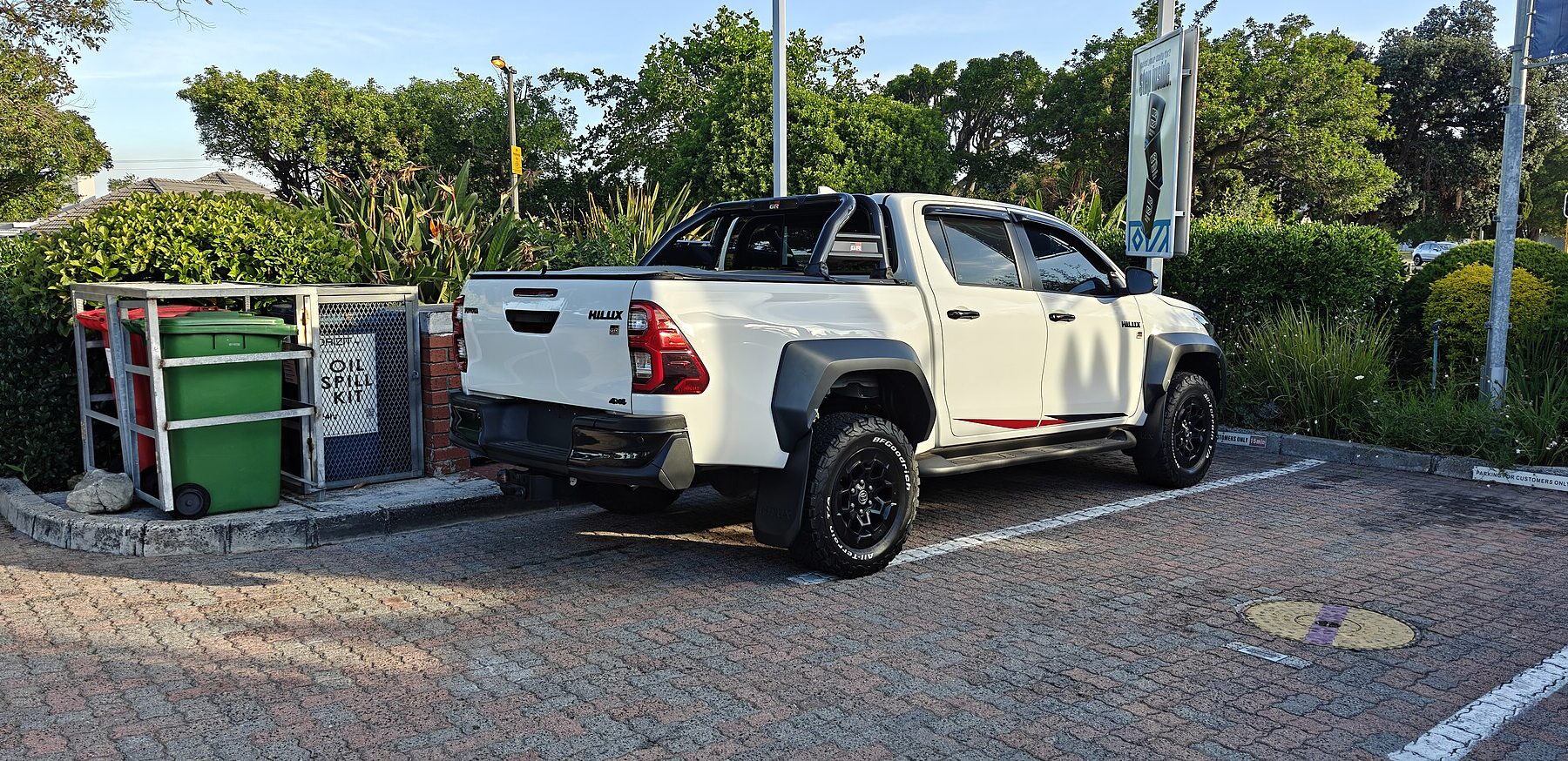 A white Toyota Hilux double-cab pickup parked in a car park beside green bushes and trees in Cape Town