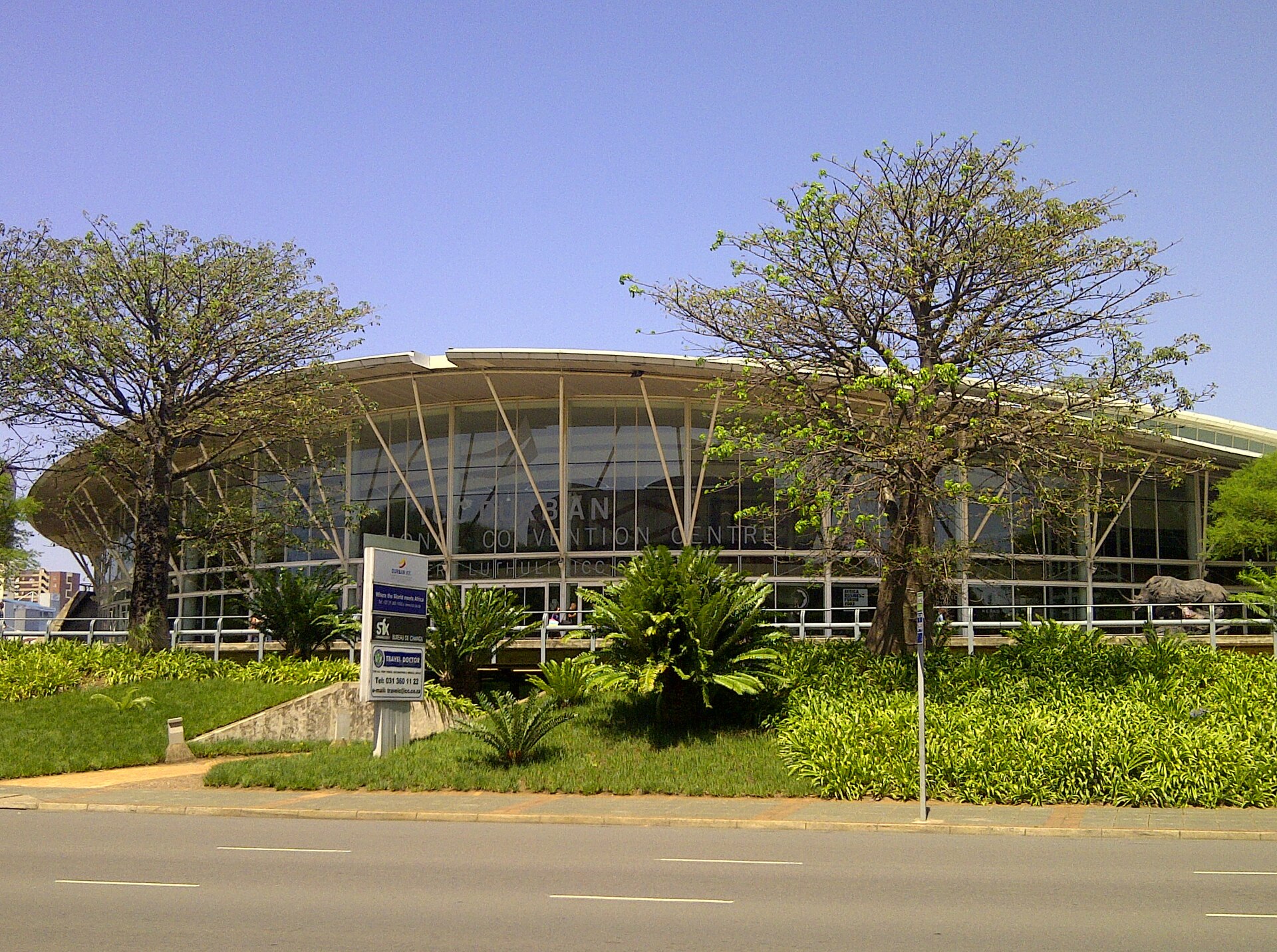 The curved glass front of the Durban International Convention Centre behind trees, palms and a road, under a clear blue sky