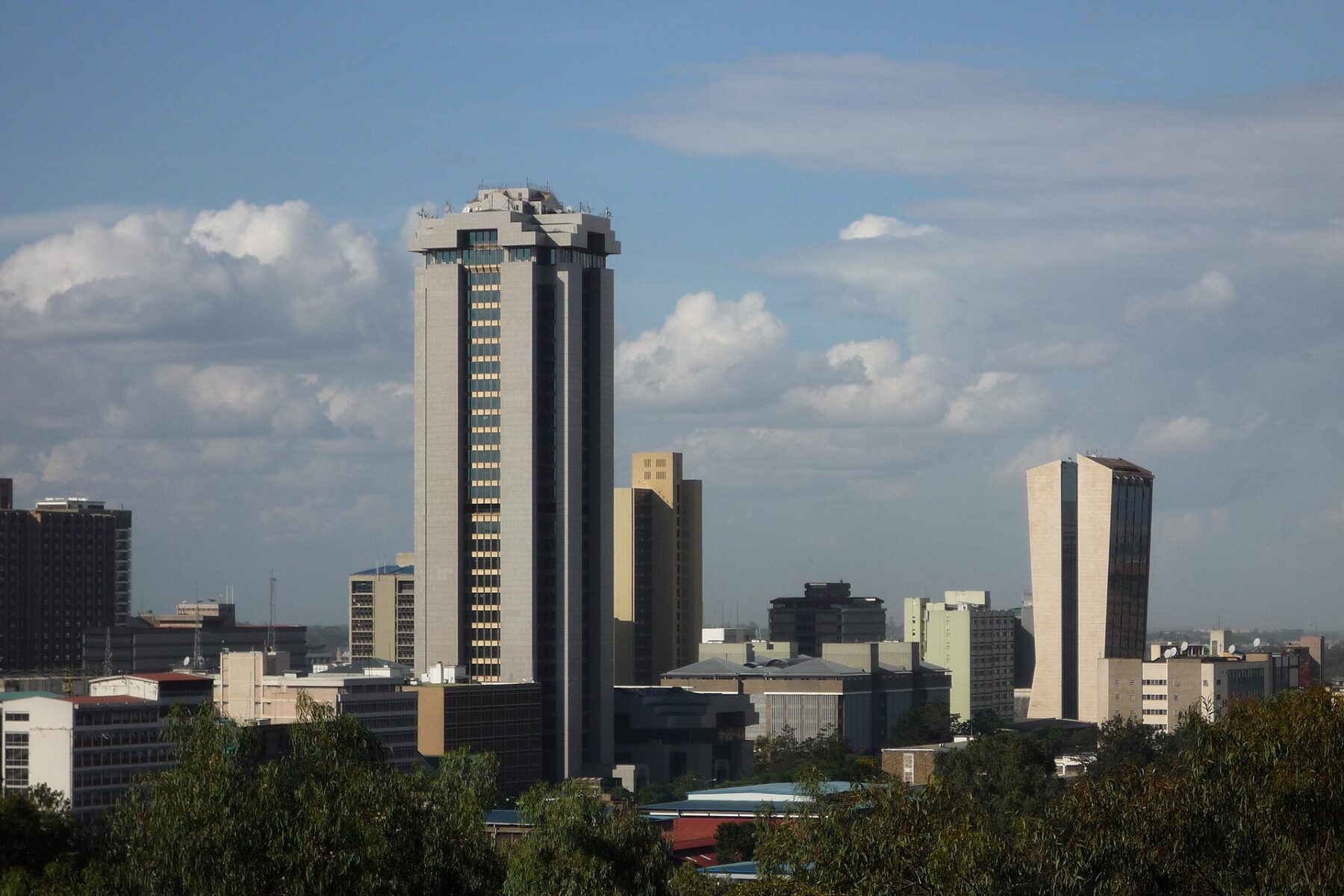 Tall office towers and lower buildings in central Nairobi behind green treetops under a blue sky with clouds