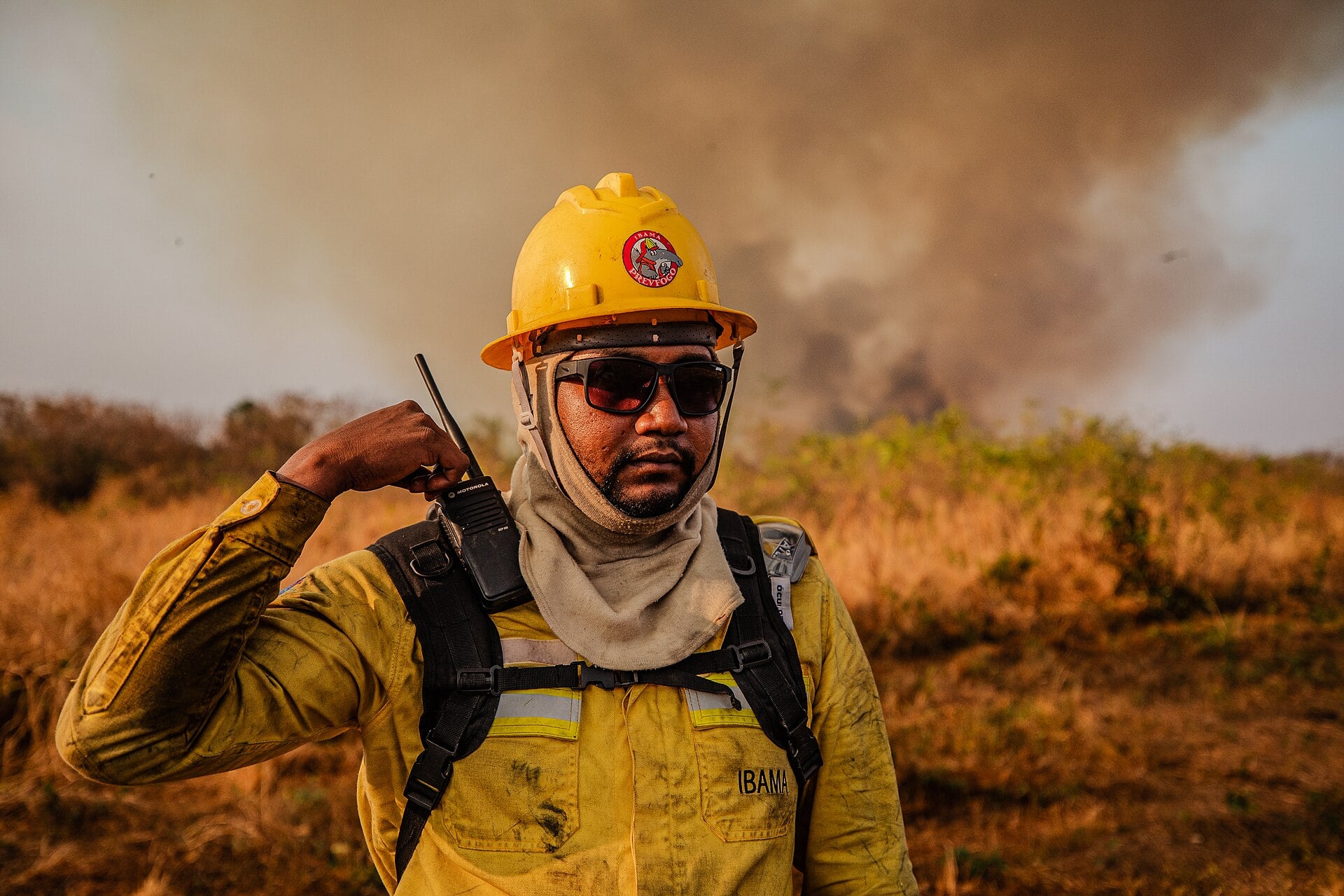 An Ibama Prevfogo firefighter in a yellow helmet and fire gear with smoke rising behind him on a burning field in Brazil