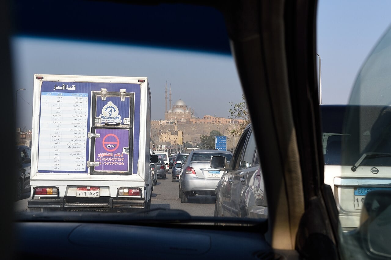 Cairo traffic seen through a car windscreen, with a delivery truck ahead and the domes and minarets of the Muhammad Ali Mosque on the Citadel in the distance.