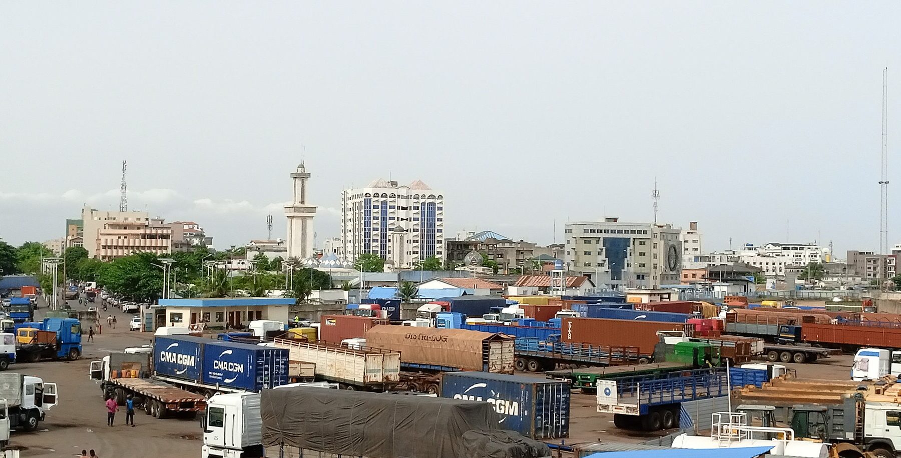 Trucks and shipping containers in the Zongo district of Cotonou, Benin, with apartment blocks and a minaret behind under an overcast sky.