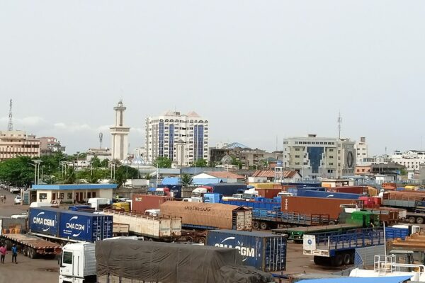 Trucks and shipping containers in the Zongo district of Cotonou, Benin, with apartment blocks and a minaret behind under an overcast sky.