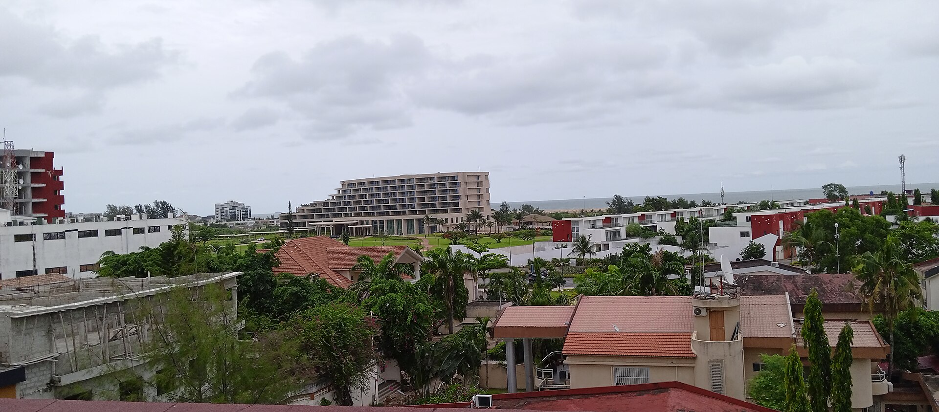 Rooftops, palm trees and a seaside hotel block with the Atlantic horizon in the Cadjehoun district of Cotonou, Benin, under grey cloud.