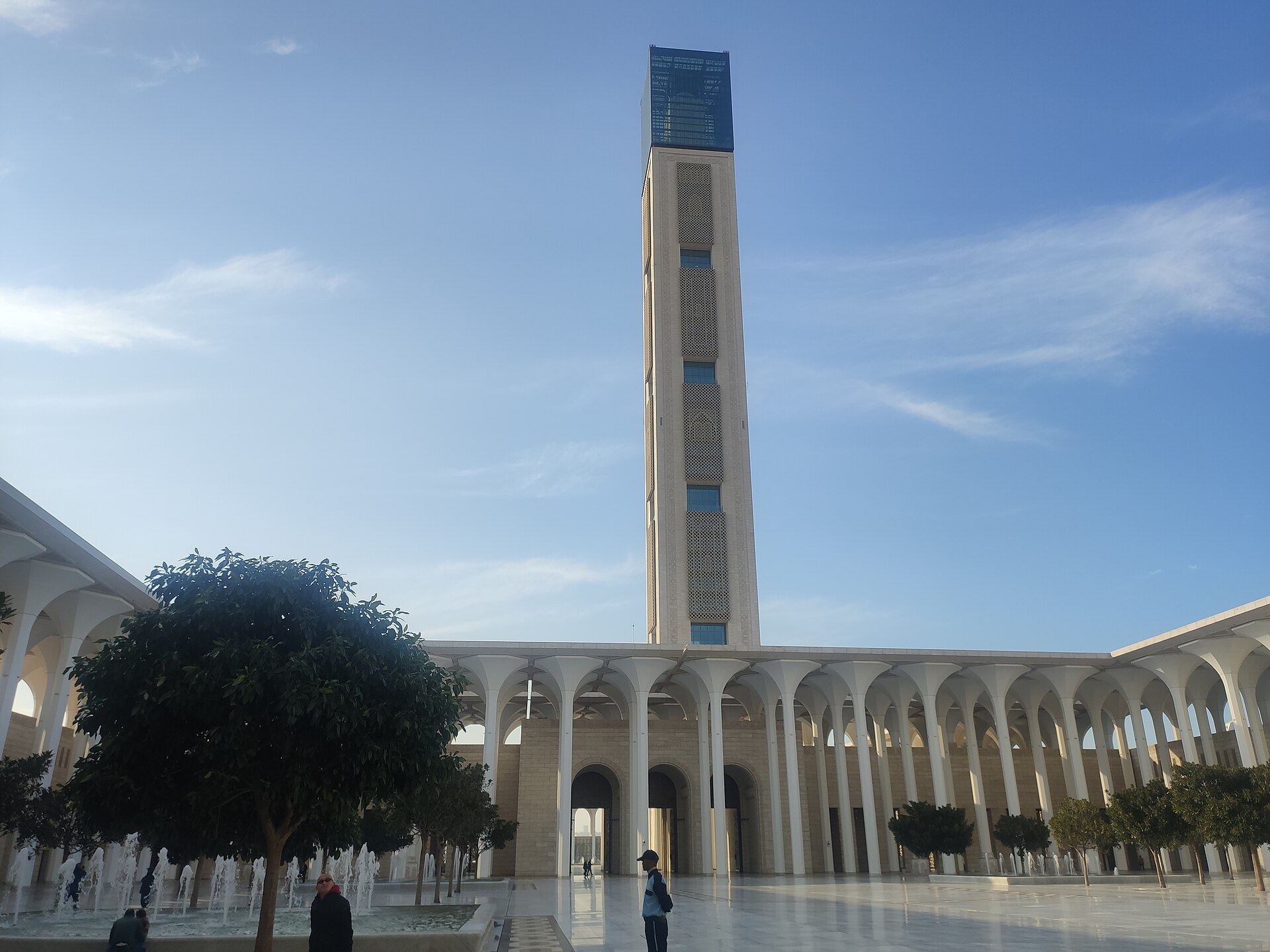 The tall minaret and white arcades of the Great Mosque of Algiers rising over a fountain courtyard under a blue sky, with a few visitors in the foreground