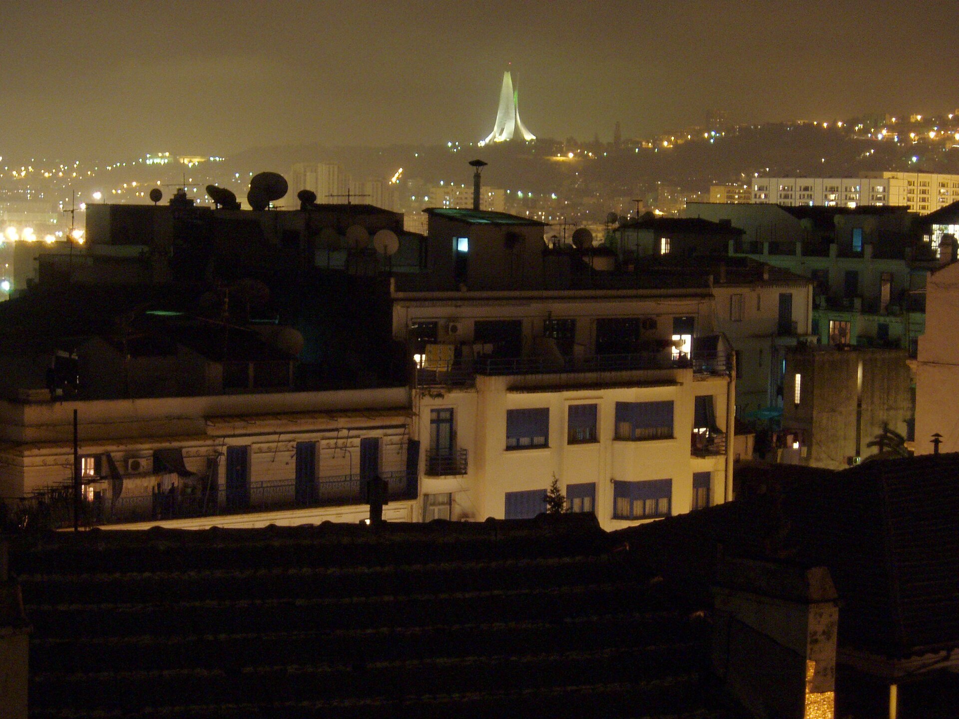 Rooftops and apartment blocks of central Algiers at night, with the illuminated Martyrs' Memorial on a hill in the background