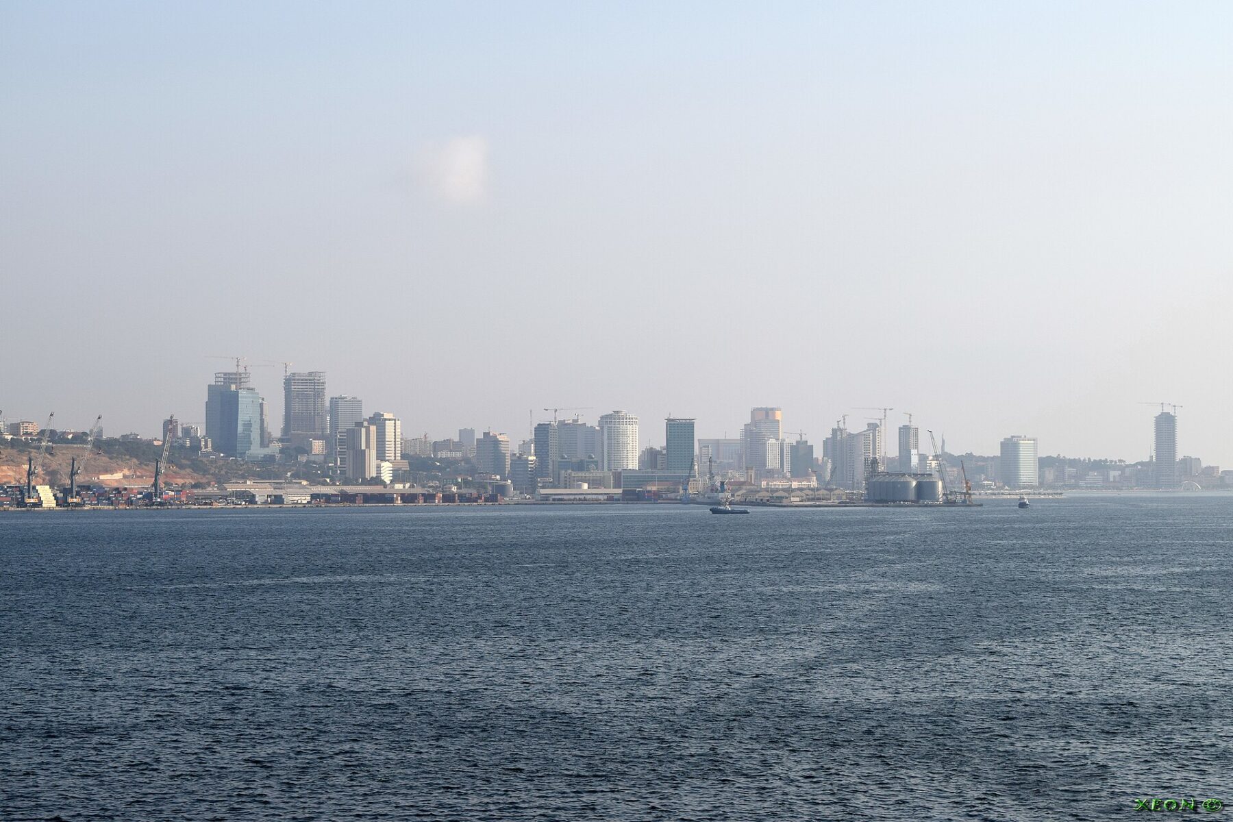 The skyline of Luanda with office towers and a port seen across the blue water of the bay under a hazy sky