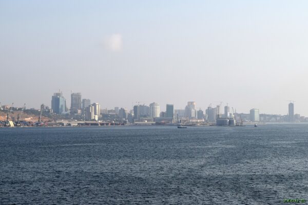 The skyline of Luanda with office towers and a port seen across the blue water of the bay under a hazy sky