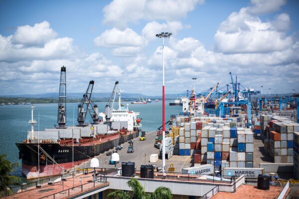 A bulk carrier moored at a quay with cranes, stacks of shipping containers and blue gantry cranes at Mombasa port under a partly cloudy sky, with the harbour behind