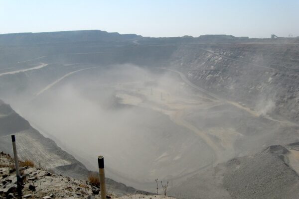 Wide open-pit mine with terraced walls and haul roads, partly hidden by a haze of dust under a pale sky