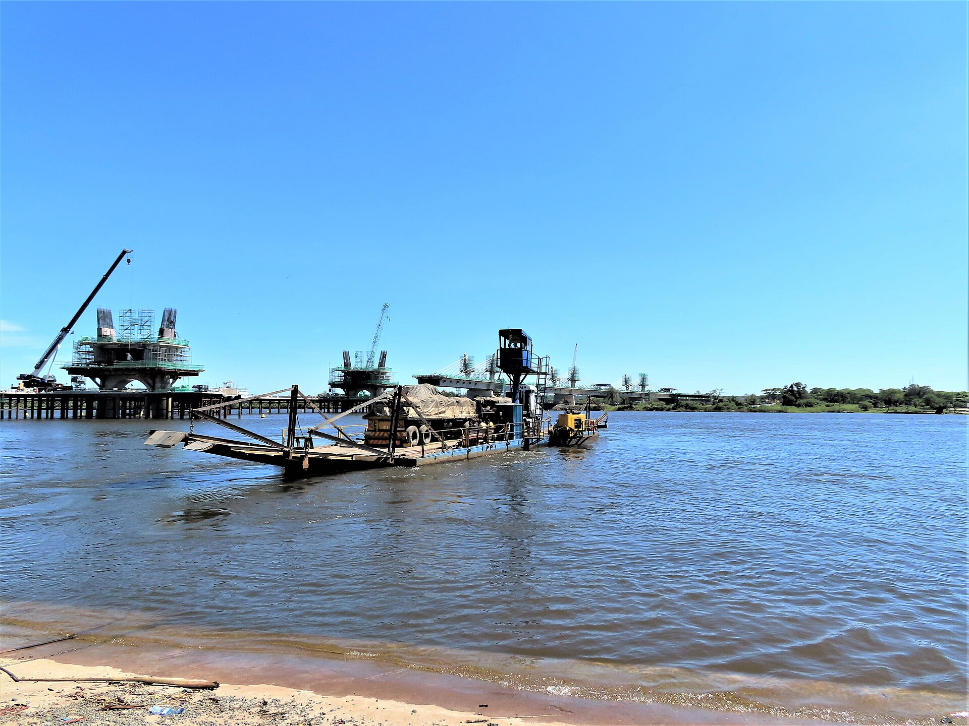 A flat truck ferry carrying a covered lorry sits on the Zambezi River beside a sandy bank, with cranes and bridge piers under construction behind