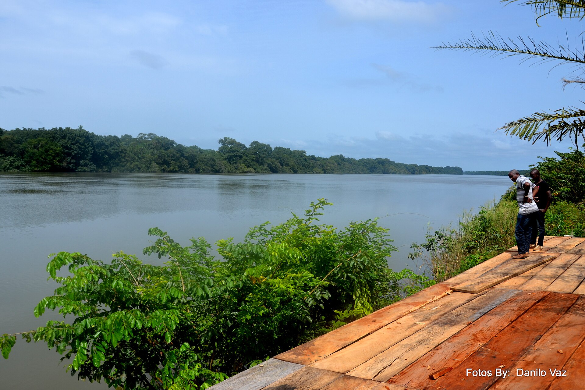 The wide, calm Corubal river at Saltinho in Guinea-Bissau, with forested banks, green shrubs and two men standing on a wooden platform at the water's edge