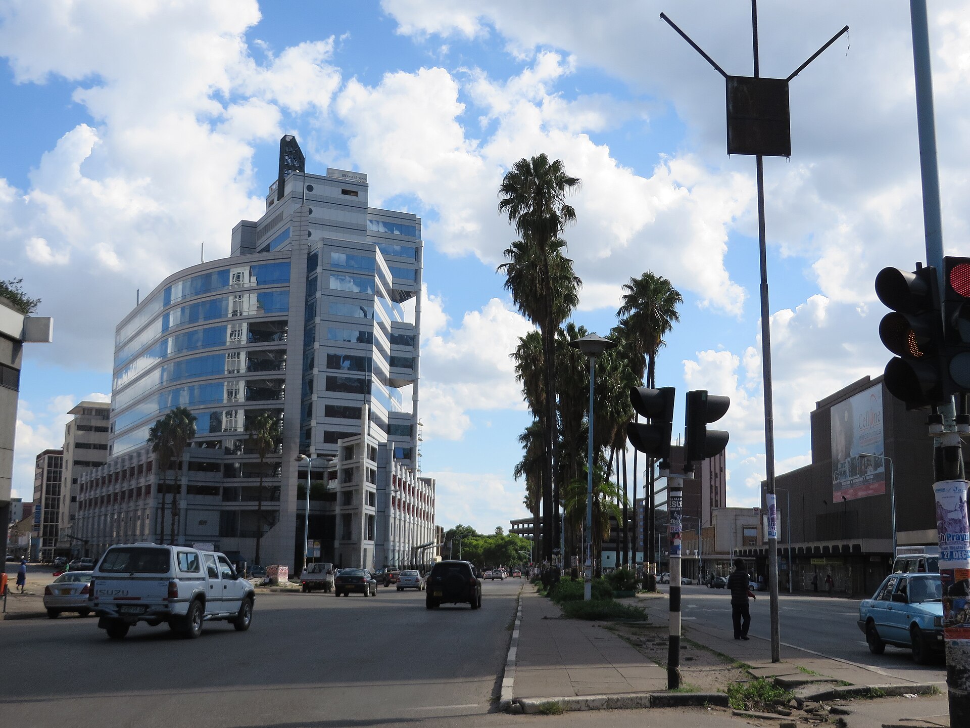 A wide avenue in downtown Harare with a curved glass office tower, palm trees, traffic lights and cars