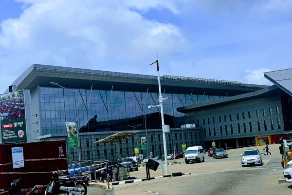 Front of the main terminal building of Nnamdi Azikiwe International Airport in Abuja with a glass facade, parked cars and a cloudy sky