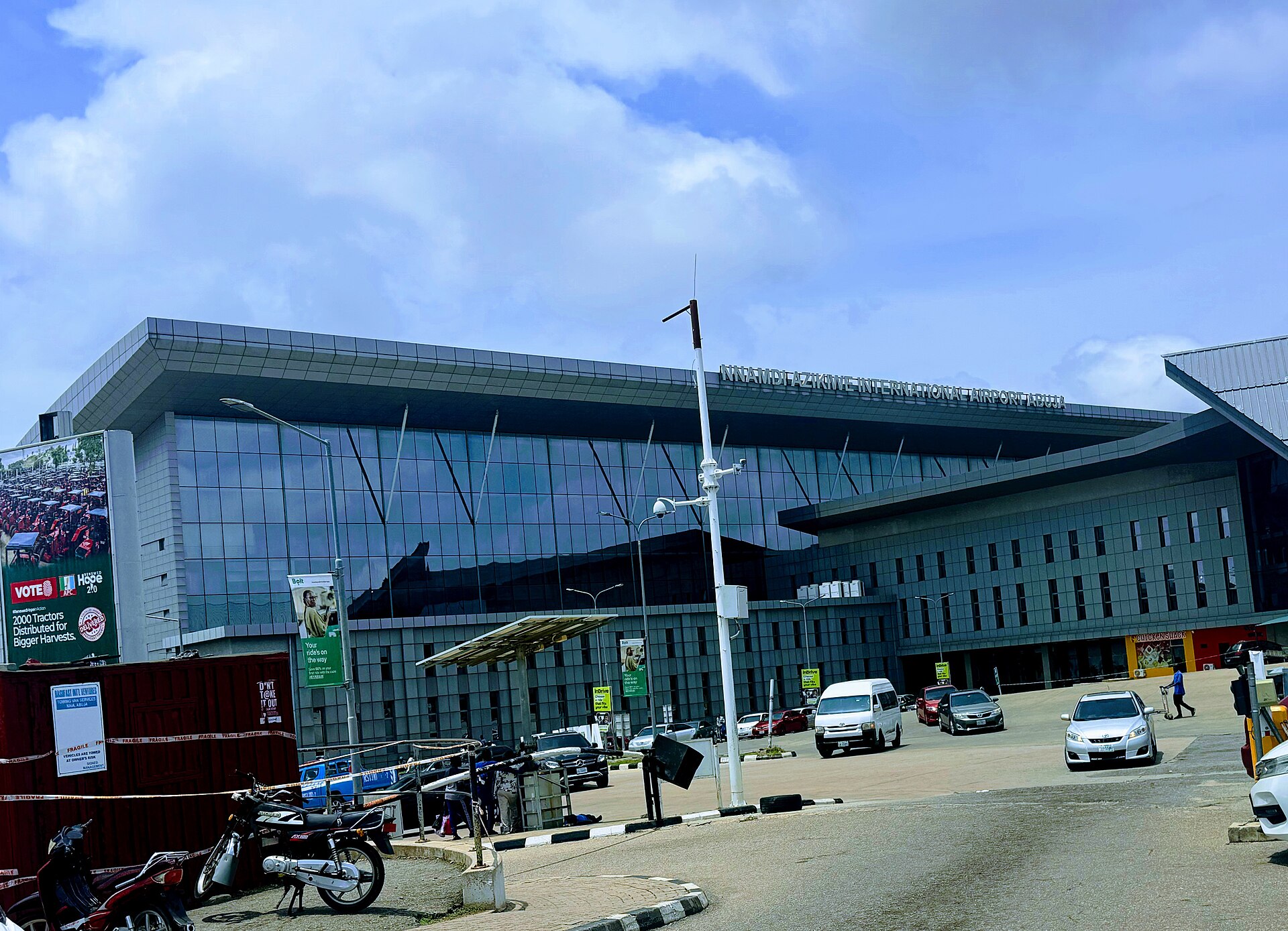 Front of the main terminal building of Nnamdi Azikiwe International Airport in Abuja with a glass facade, parked cars and a cloudy sky