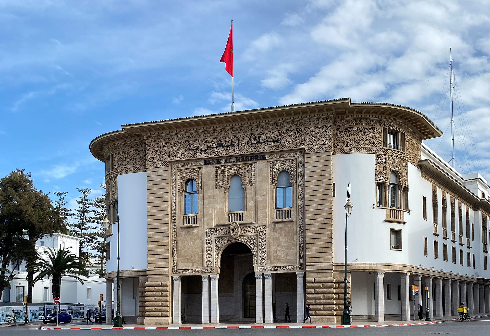 Sand-coloured facade of the Bank Al-Maghrib building in Rabat with carved stonework, arched windows and a Moroccan flag on the roof, under a blue sky