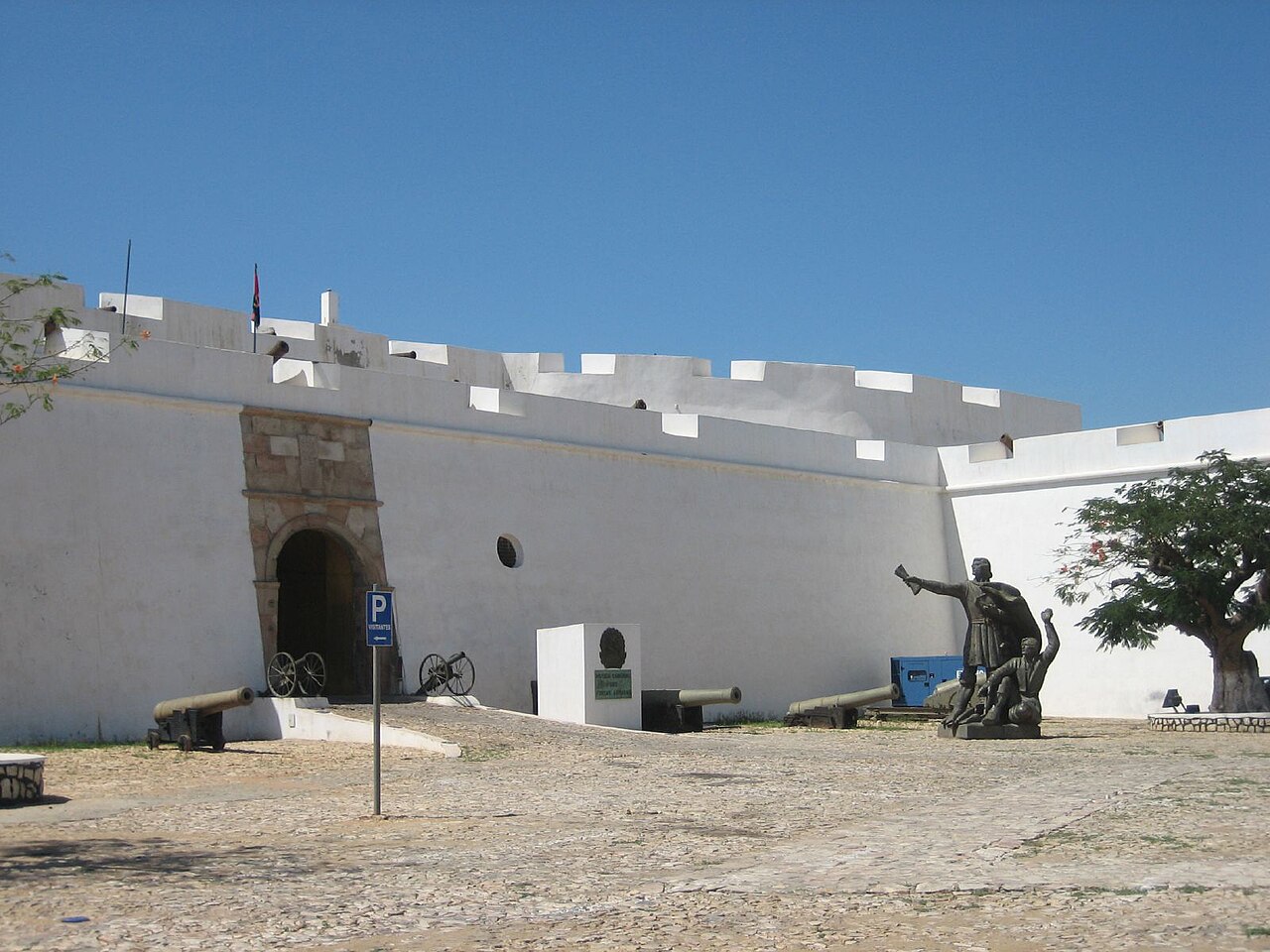 A whitewashed fort wall with an arched entrance, old cannons and a bronze statue group on a cobbled square in Luanda.