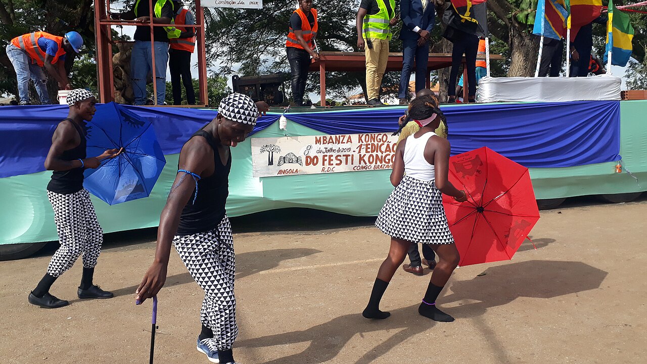 Two dancers in black-and-white patterned outfits and a woman holding a red umbrella move on a dirt road in front of a parade float with a banner reading Mbanza Kongo.