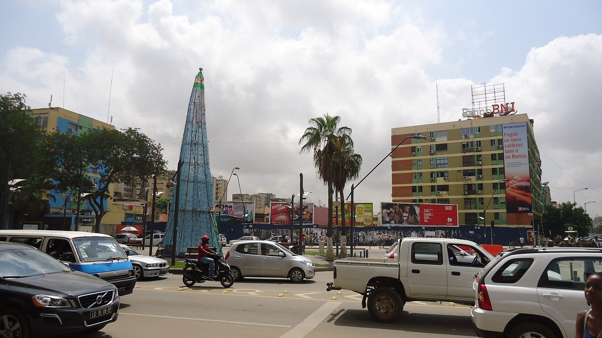 Traffic and apartment blocks on an avenue in central Luanda with billboards and a tall decorative tree