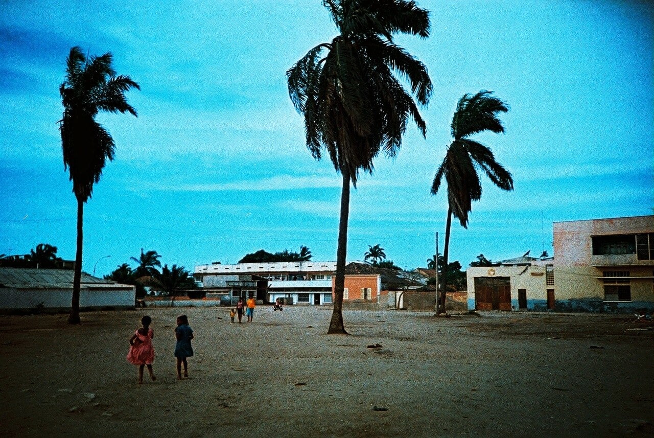 Children walk across a sandy square lined with tall palm trees and low colonial buildings in Benguela, Angola, under a deep blue sky