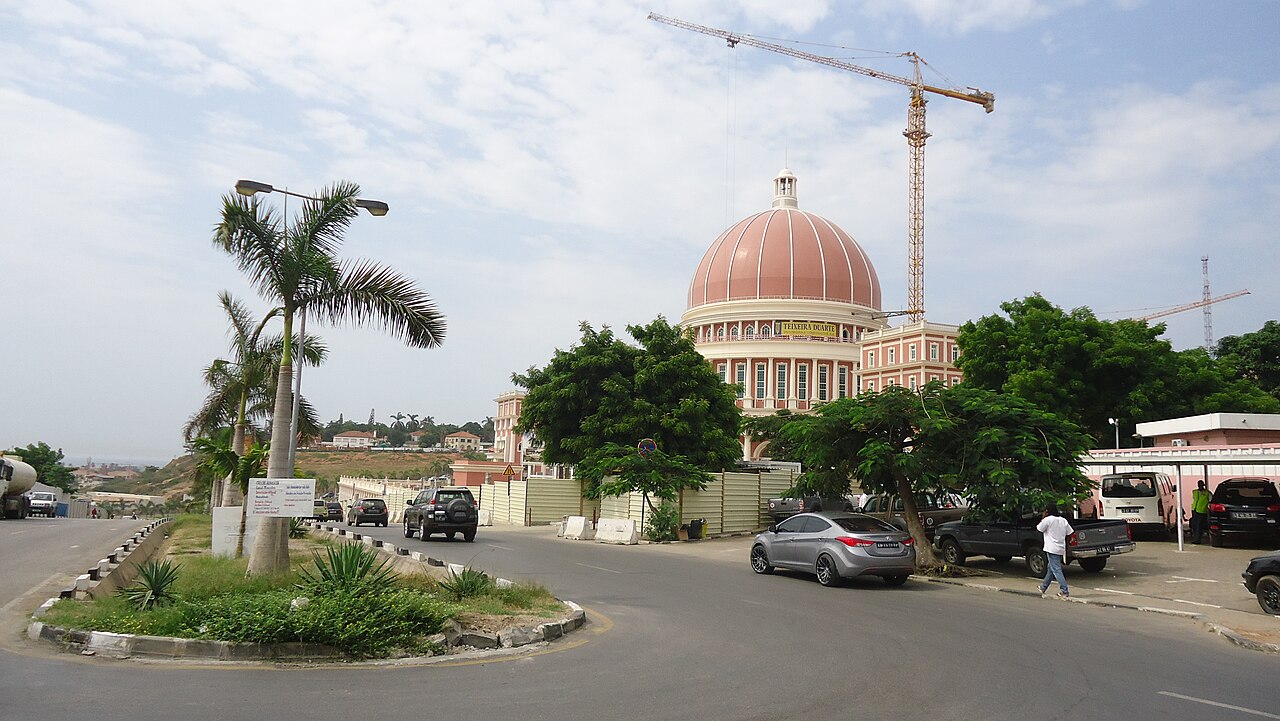 The domed new National Assembly building under construction beside a road in Luanda, with palm trees and a crane