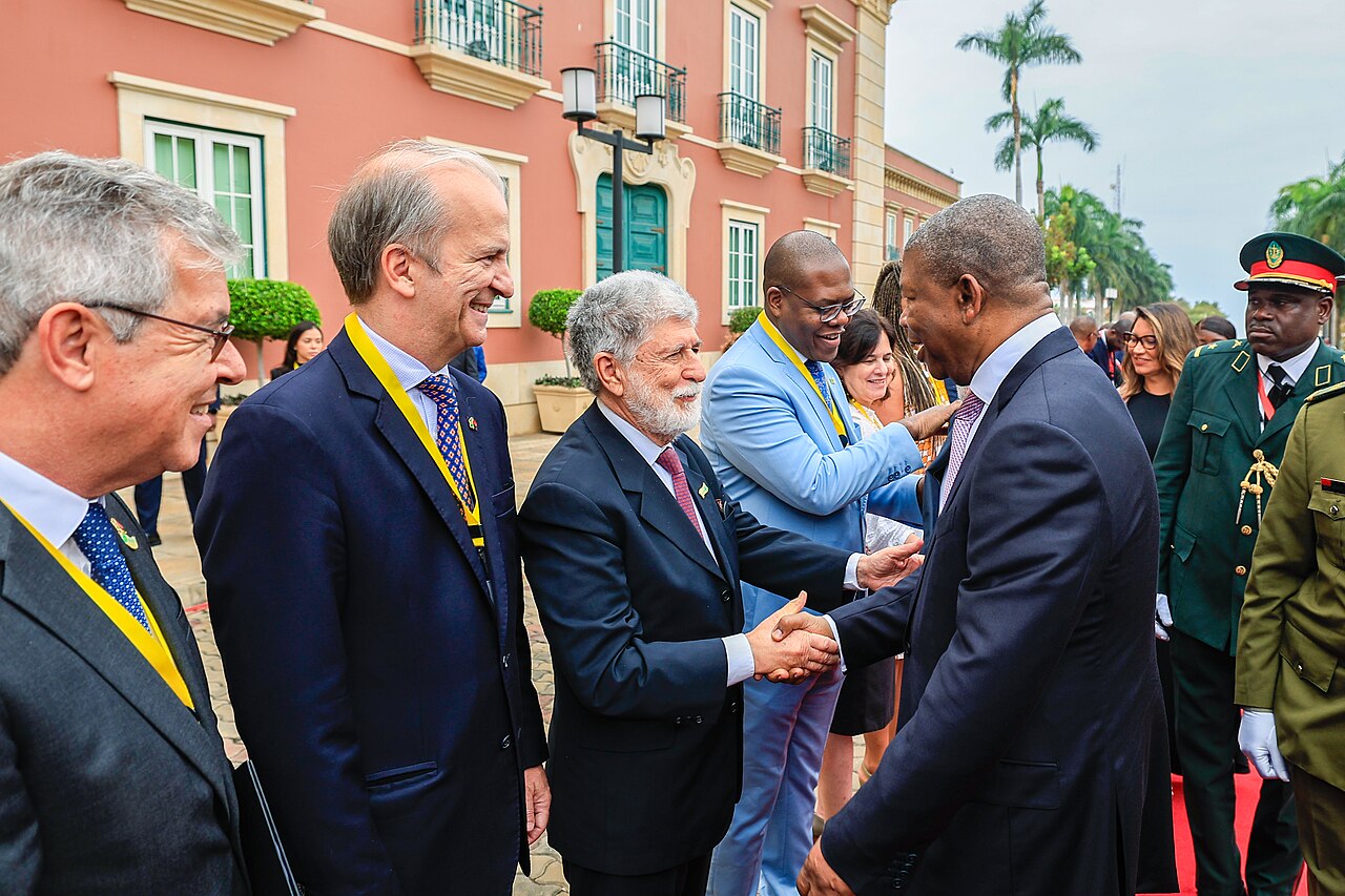 Officials in dark suits and uniformed guards greeting each other outside the pink Presidential Palace in Luanda
