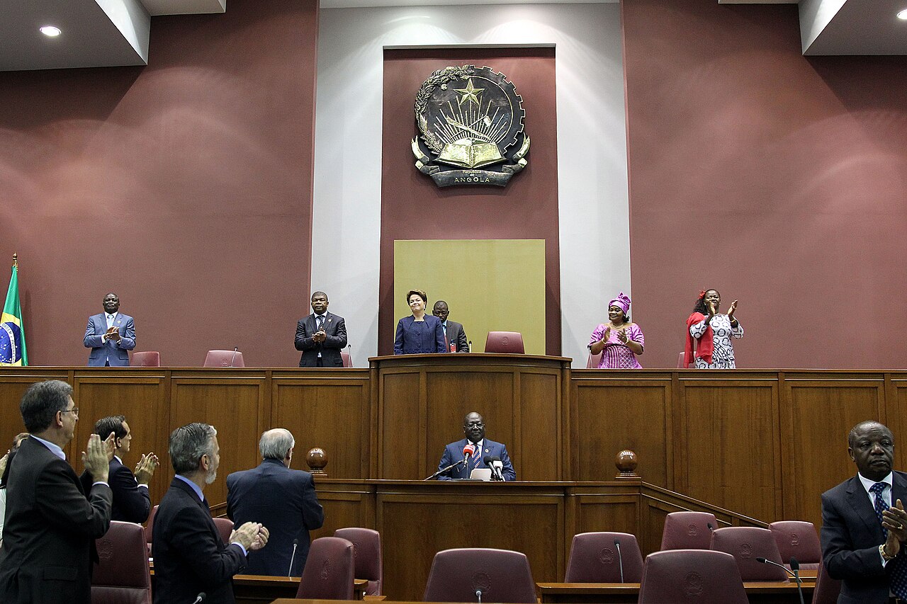 Lawmakers and guests stand and applaud in the chamber of Angola's National Assembly beneath the national coat of arms