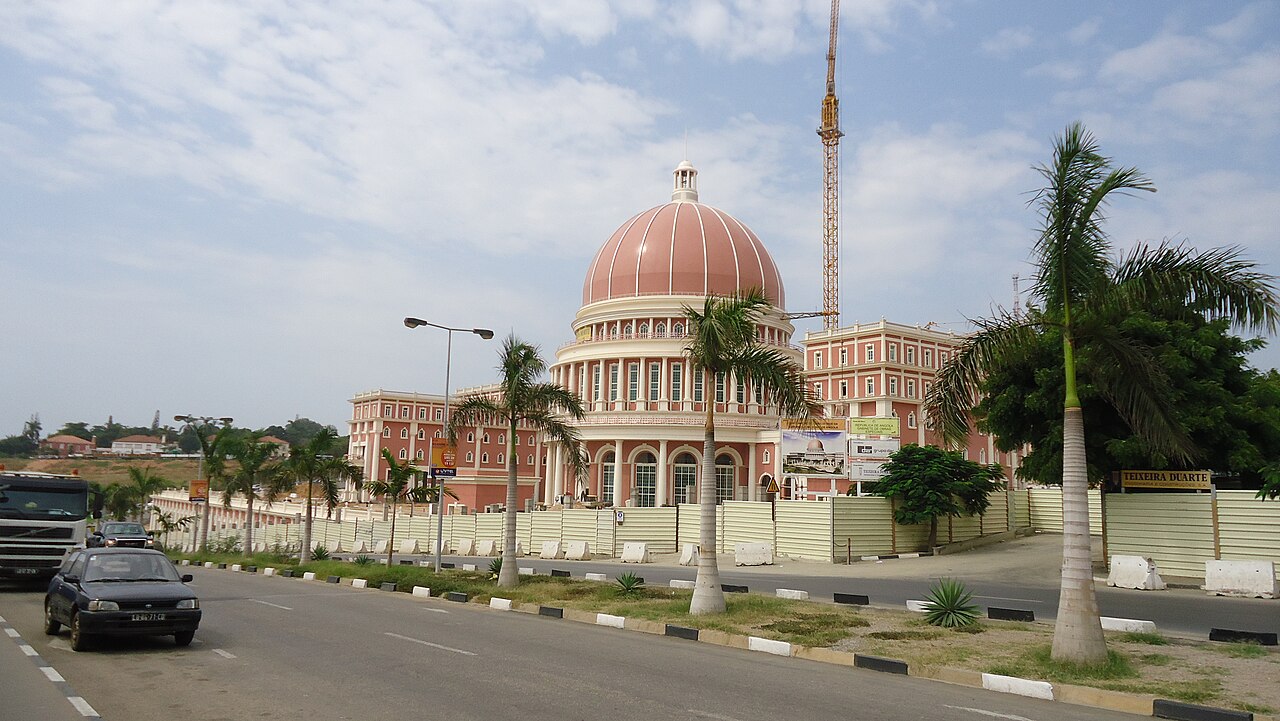 Pink domed National Assembly building in Luanda behind palm trees and a construction fence, with a crane beside the dome