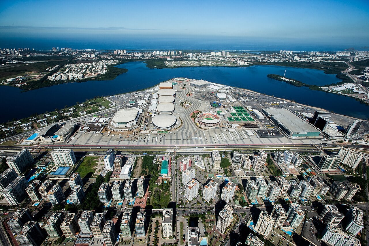 Aerial view of the Olympic Park in Barra da Tijuca, Rio de Janeiro, with the arenas and stadium beside a lagoon and apartment towers across the road.