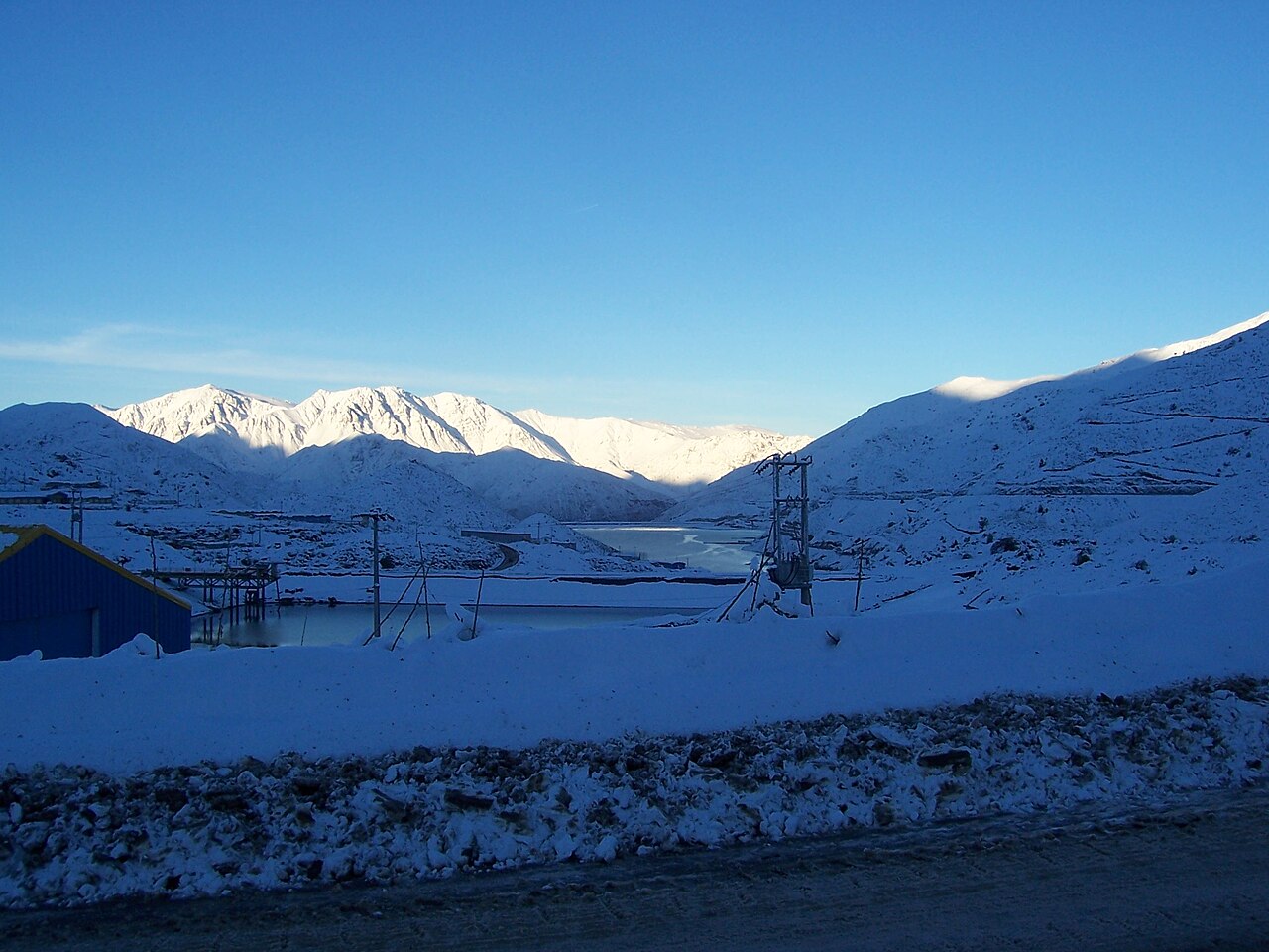 A frozen reservoir and snow-covered mountains at the Los Pelambres mine in Chile, with a blue industrial shed at left