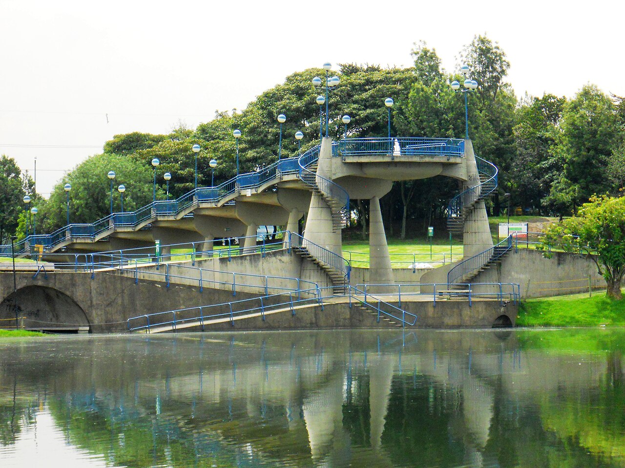 A concrete walkway on tall pillars with blue railings and rows of globe lamps beside a lake in a park, with green trees behind.