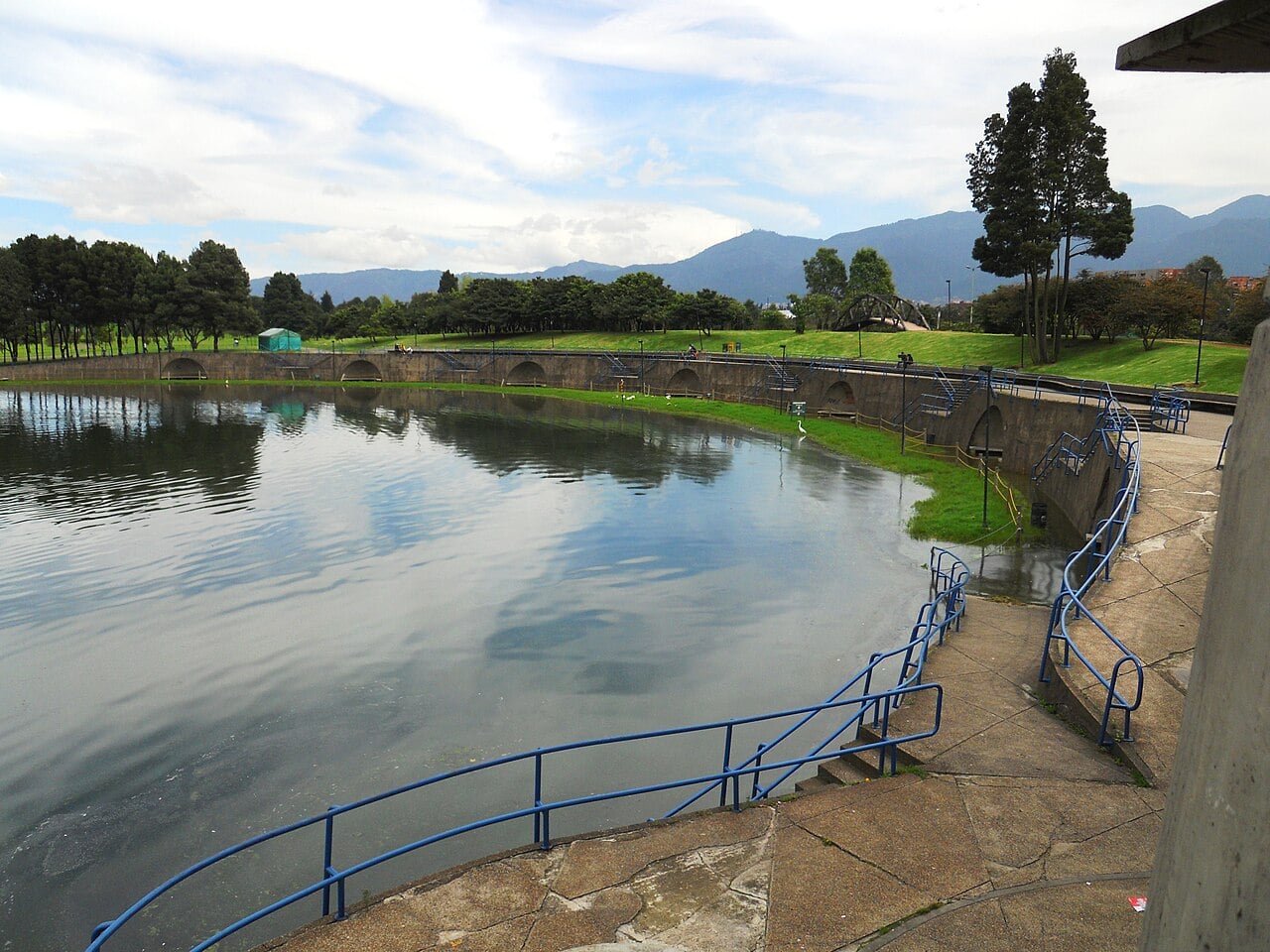 A lake with a curved stone embankment and blue railings in a green park, with mountains and cloud behind, in Parque Simón Bolívar, Bogotá.