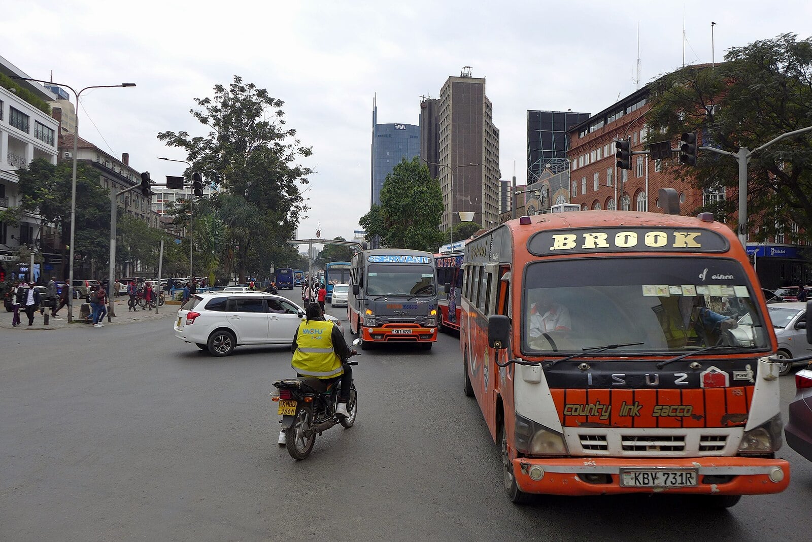 Matatu minibuses and a motorcyclist in traffic on Kenyatta Avenue in central Nairobi, with office towers behind