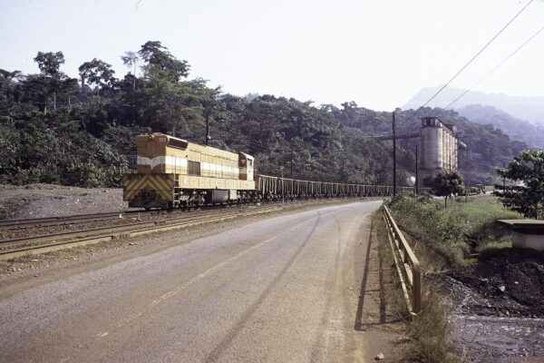 Locomotive number 303 pulling a long train of iron ore wagons past loading silos in forest country at Yekepa, Liberia