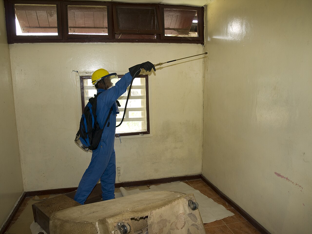 A worker in blue overalls, a yellow helmet and a face shield sprays insecticide on the wall of a room in Yekepa, Liberia