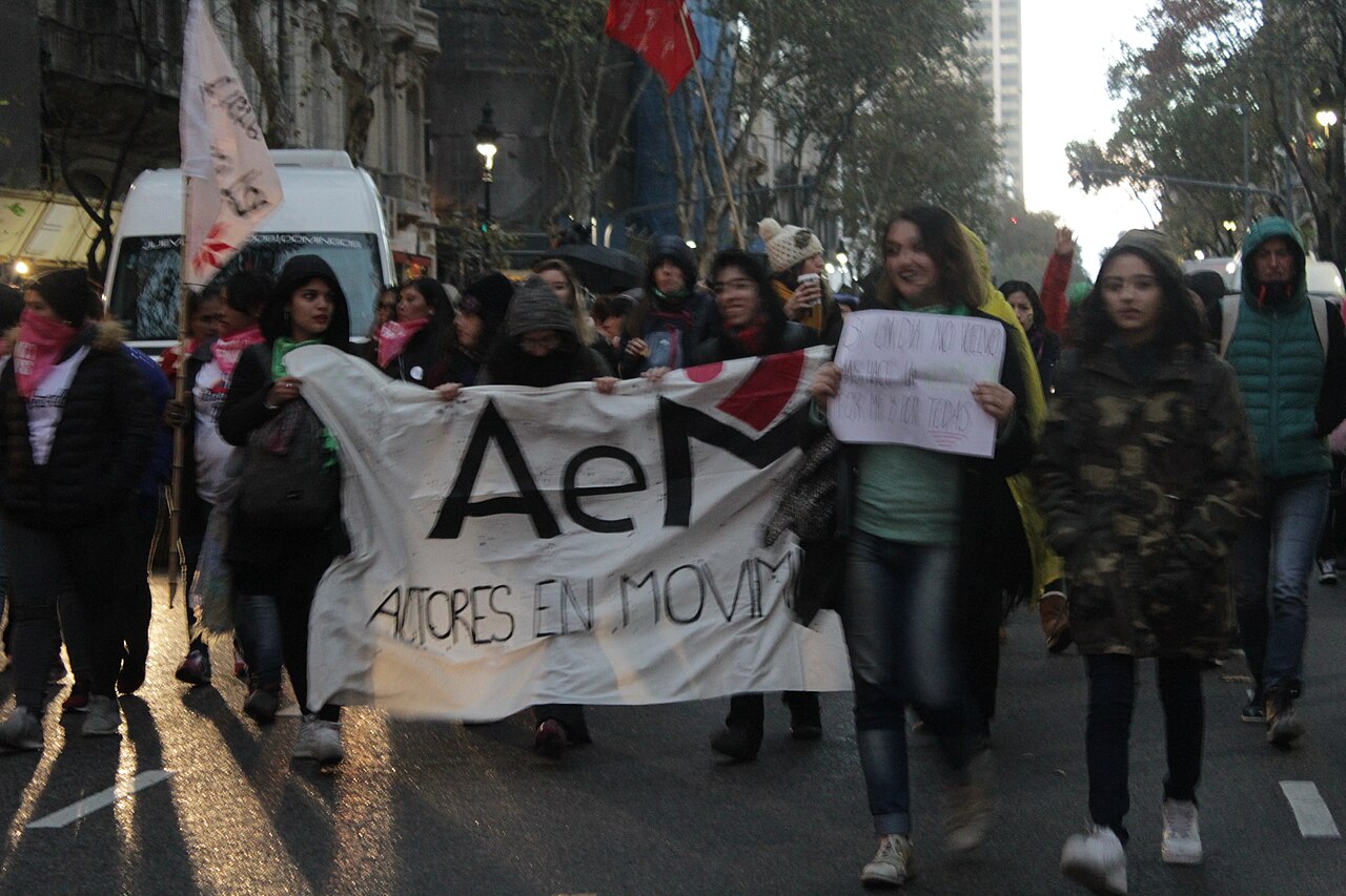 Women march down a Buenos Aires avenue behind a white banner, one holding a handwritten sign, with flags overhead