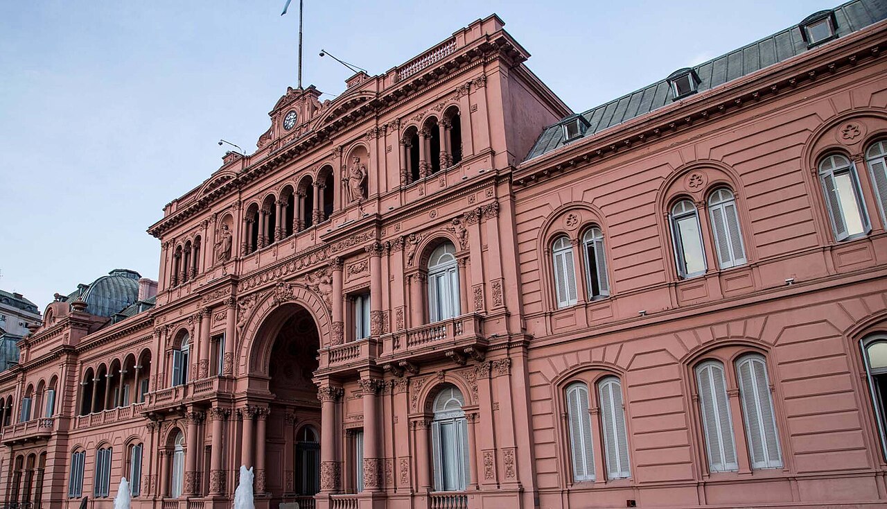The pink facade of the Casa Rosada presidential palace in Buenos Aires, with arched windows, balconies and a clock under a pale sky