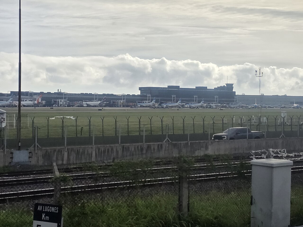 Terminal and parked airliners at Aeroparque Jorge Newbery seen across a fence and railway tracks on Avenida Lugones