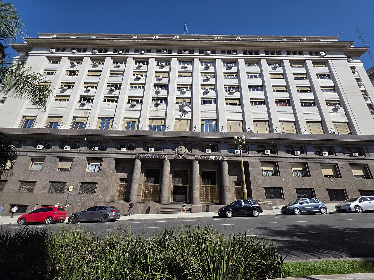 The stone and granite facade of Argentina's Ministry of Economy building in Buenos Aires, with Argentine flags on the roof and the upper facade.