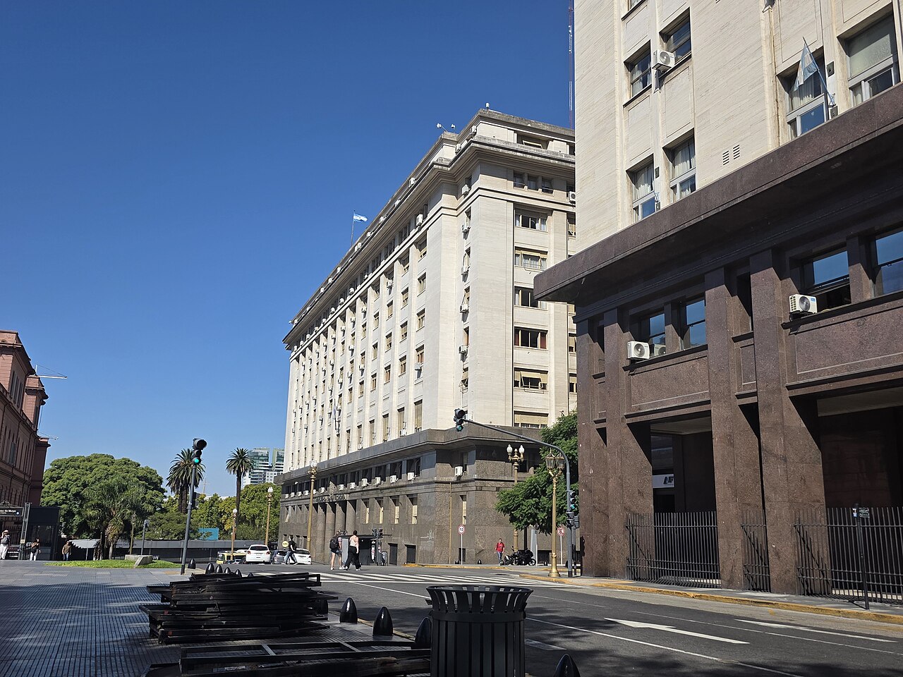 The white stone Economy Ministry building on Hipólito Yrigoyen street in Buenos Aires, with the pink Casa Rosada and palm trees at left and a granite-fronted office block at right