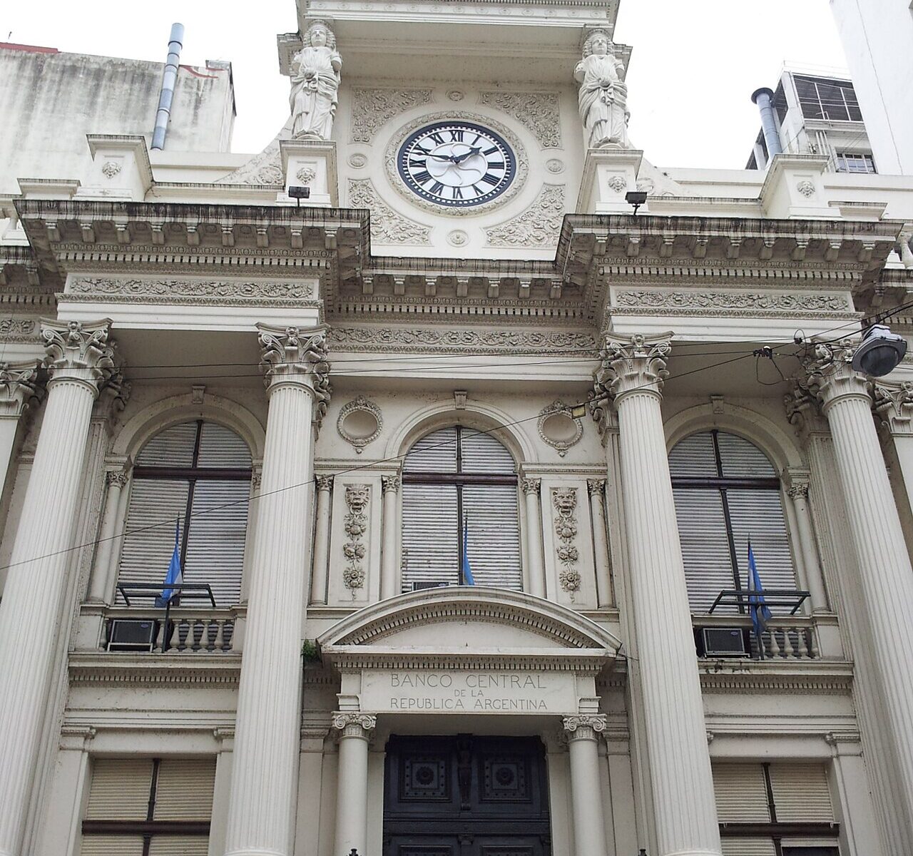 Columned white facade of Argentina's central bank in Buenos Aires, with a clock, statues and flags above a dark wooden door