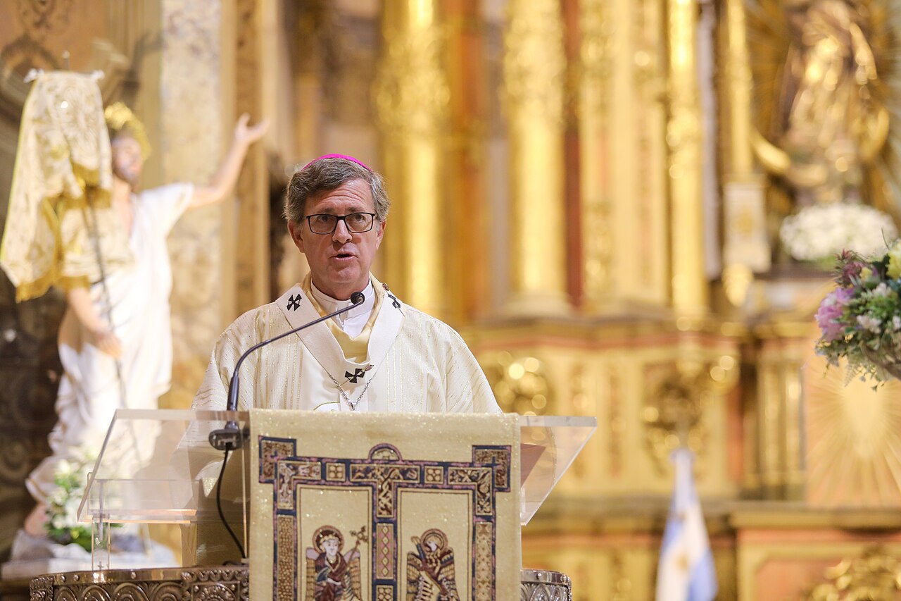Archbishop Jorge García Cuerva in white vestments and glasses speaks at a lectern inside a gilded church