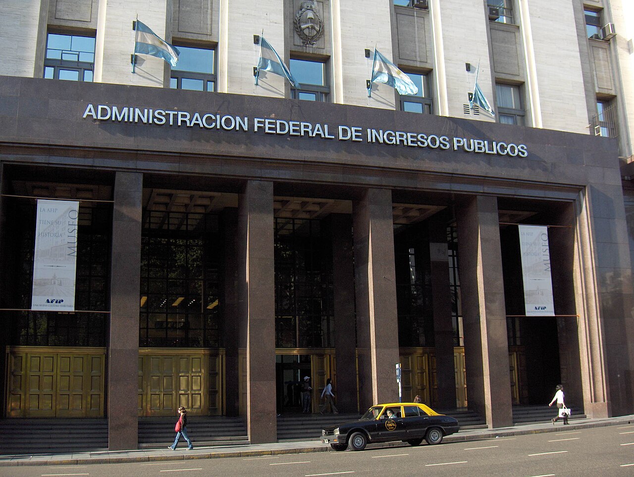 The granite entrance of Argentina's federal tax agency building in Buenos Aires, with Argentine flags above and a black and yellow taxi in front