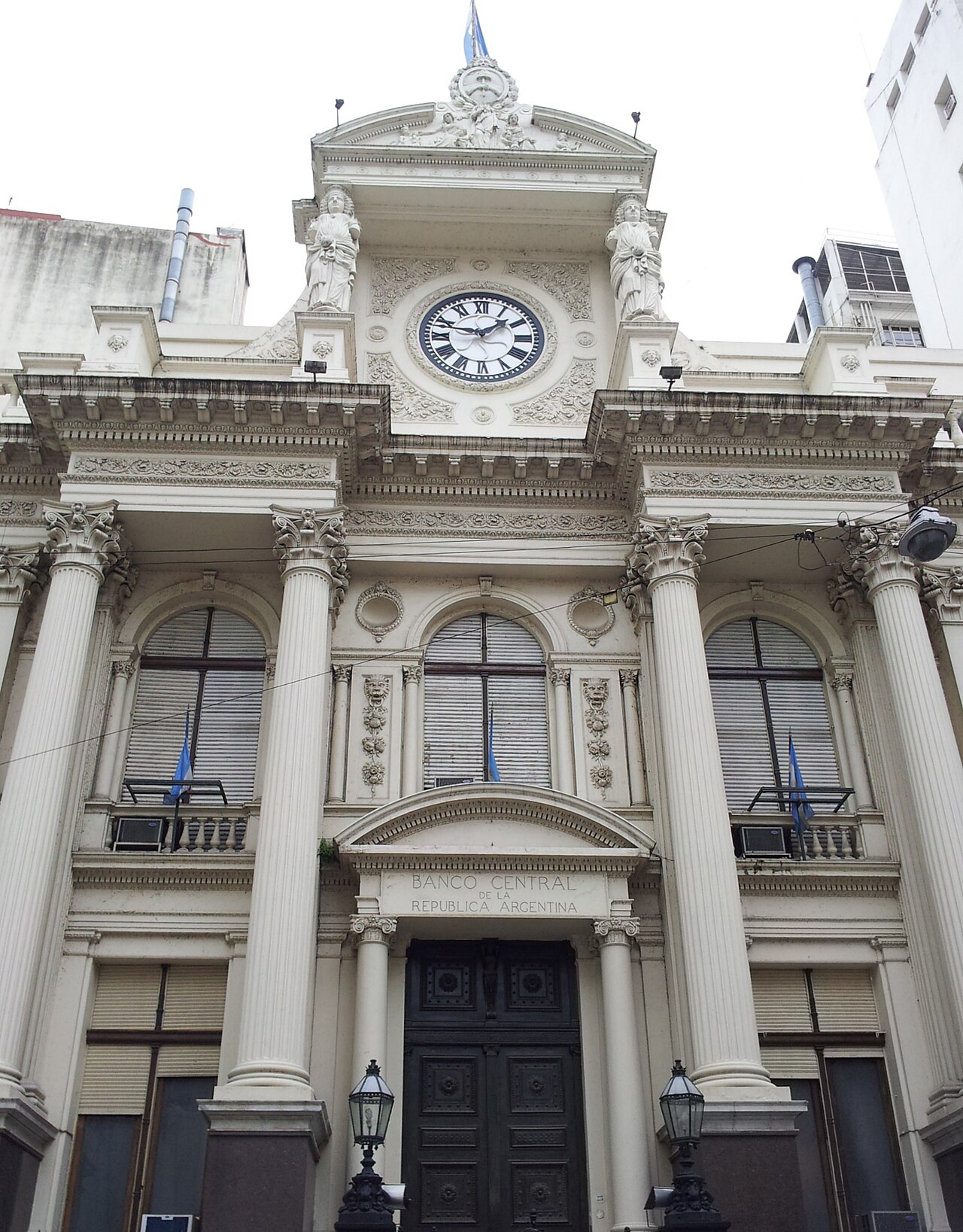Columned white facade of the Banco Central de la República Argentina in Buenos Aires, with a clock, statues and Argentine flags on the balconies above the dark wooden door