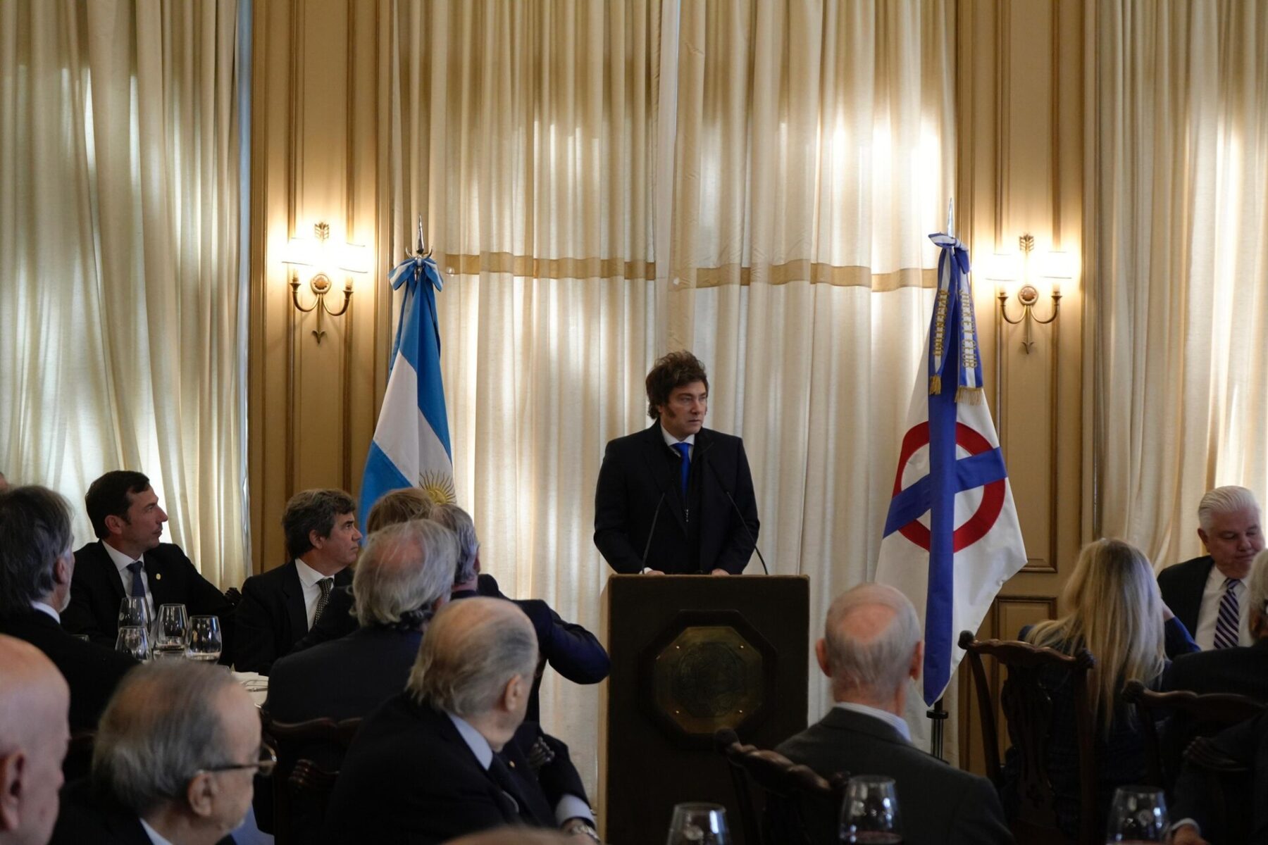 Javier Milei speaks at a lectern between the Argentine flag and a club banner