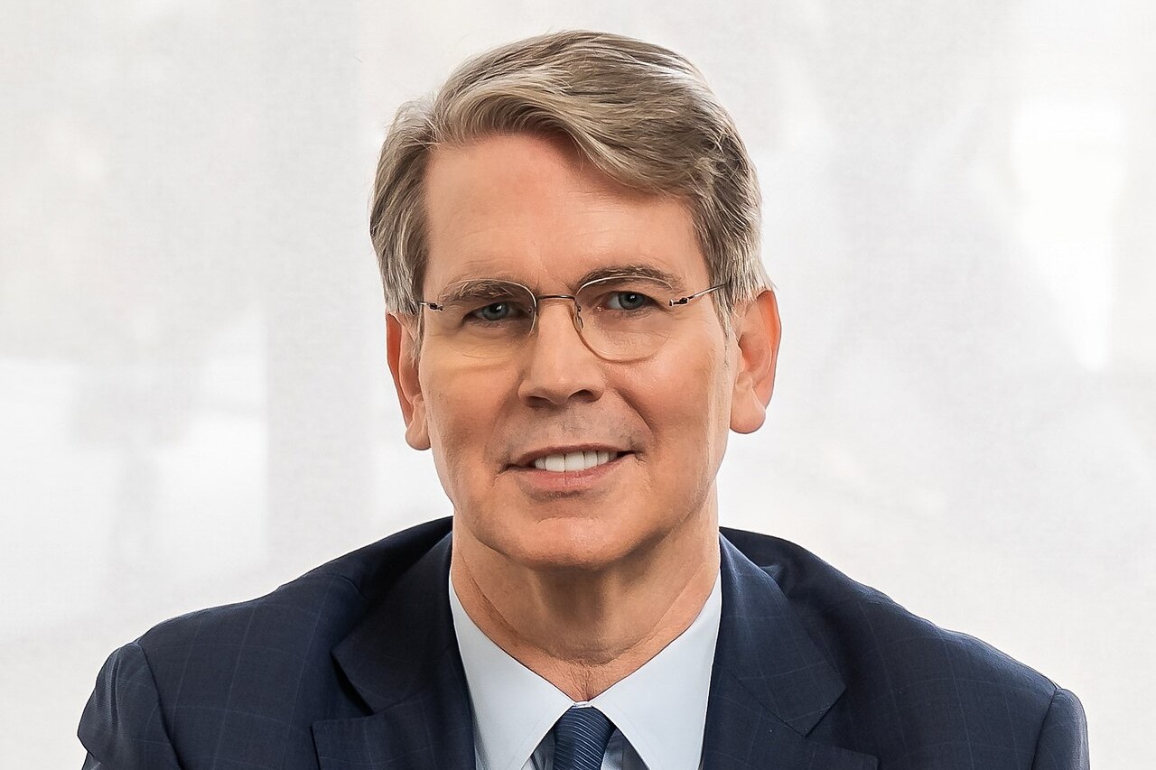 Scott Bessent, wearing glasses, a navy suit and a blue tie, smiles in front of a pale background